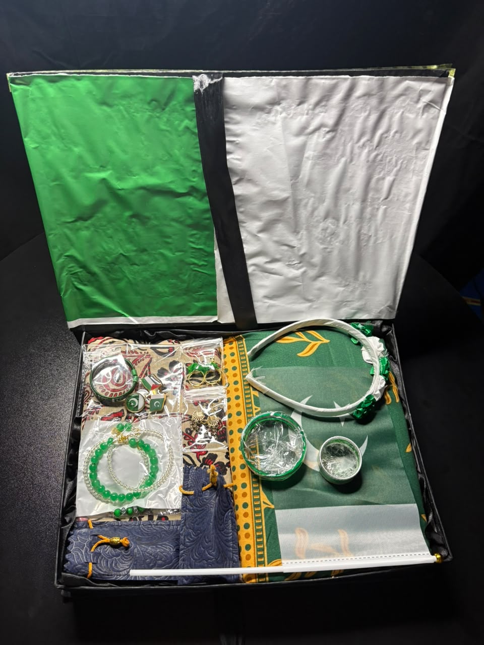 An open gift box containing a green and white headband, several green beaded bracelets, a small green and white cup, and other accessories arranged on a dark background.
