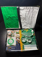 An open gift box containing a green and white headband, several green beaded bracelets, a small green and white cup, and other accessories arranged on a dark background.