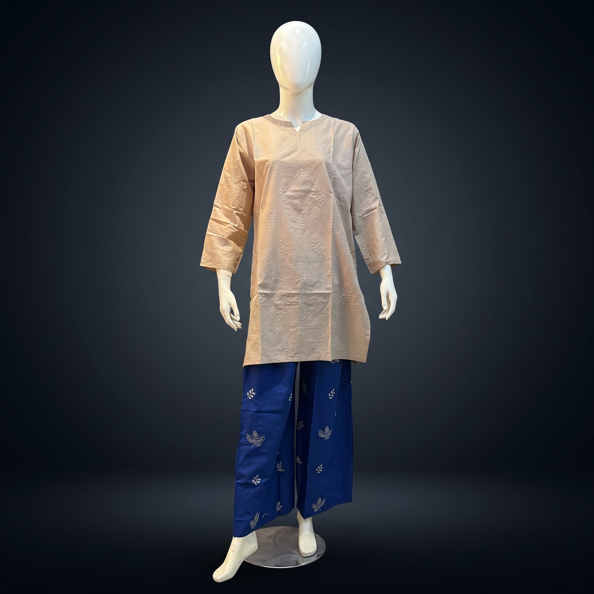 A two-piece traditional outfit displayed on a white mannequin against a dark studio background. The set includes a light beige long-sleeve tunic with a mandarin collar and a matching beige waist tie, paired with wide-leg royal blue pants featuring small silver motifs.