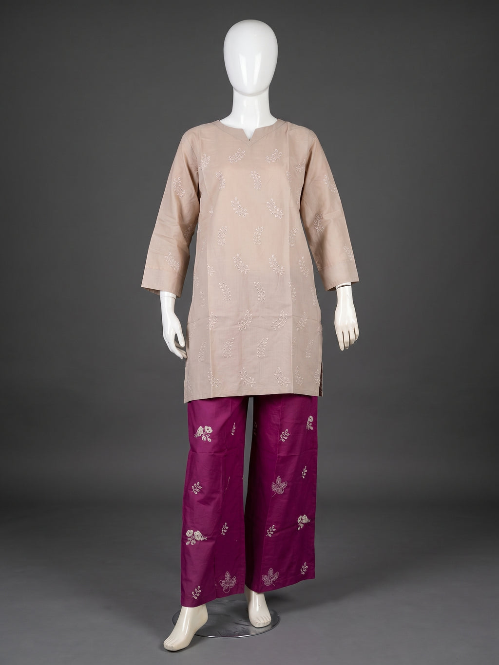 A traditional outfit displayed on a white mannequin against a dark gray studio background. The outfit consists of a light beige tunic with subtle silver embroidery and magenta wide-leg pants with silver floral motifs.