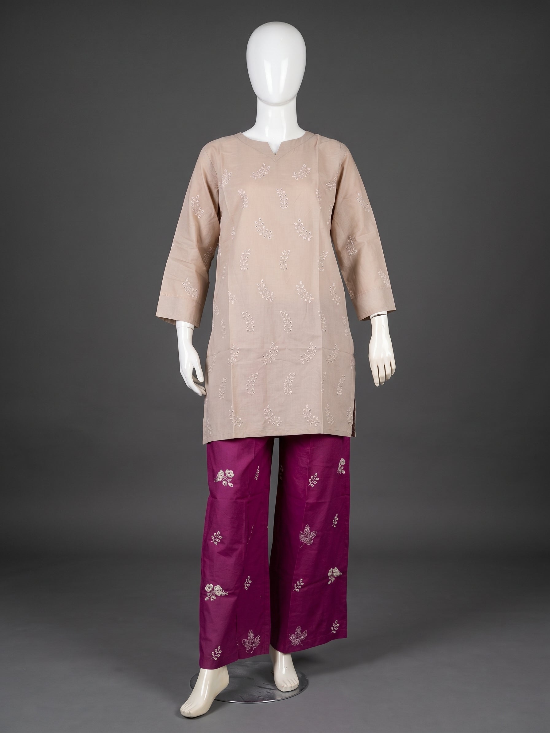 A traditional outfit displayed on a white mannequin against a dark gray studio background. The outfit consists of a light beige tunic with subtle silver embroidery and magenta wide-leg pants with silver floral motifs.