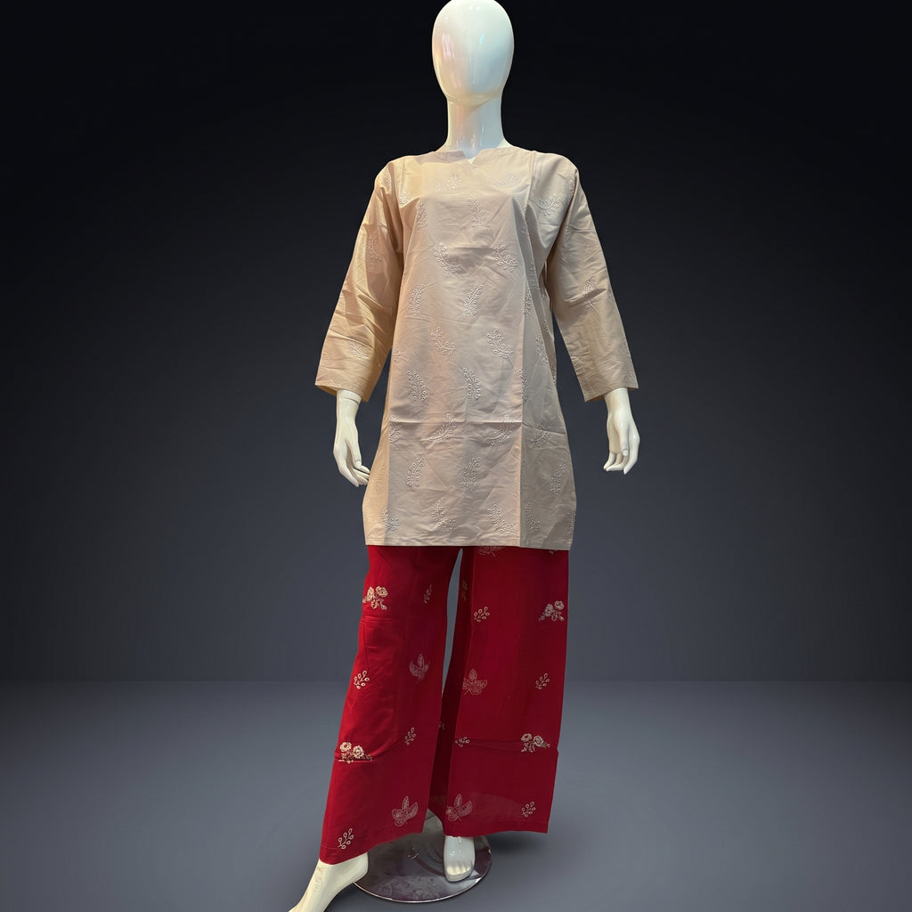 A mannequin wearing a loose beige tunic top with three-quarter sleeves and red wide-leg pants decorated with small gold floral motifs, set against a dark gray studio background.