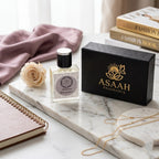 ASAAH Fragrance Maison Elegance Eau De Parfum bottle standing next to its textured black and gold presentation box on a marble slab with a single cream rose.
