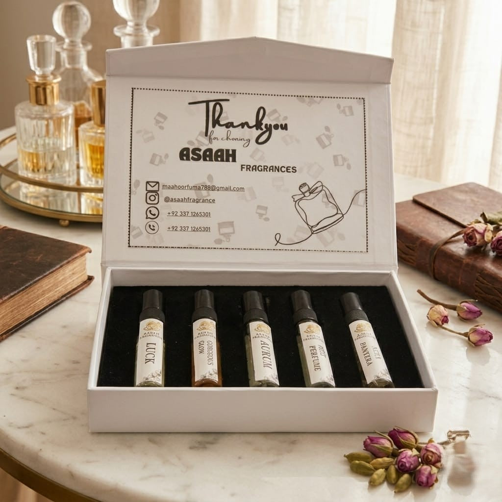 Five glass sample perfume atomizers from ASAAH Fragrance standing in a row on a wooden table in front of stacked white and gold gift boxes and a vintage clock.