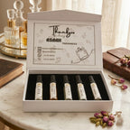 Five glass sample perfume atomizers from ASAAH Fragrance standing in a row on a wooden table in front of stacked white and gold gift boxes and a vintage clock.