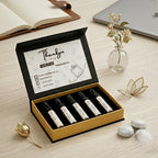 Open ASAAH Fragrance gold and black tester box on a wooden desk displaying five sample spray vials including Legend and Hafsa next to white jasmine flowers.